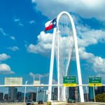 Iconic view of Margaret Hunt Hill Bridge with Texas flag and Dallas skyline under a blue sky.